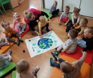 Grupa dzieci siedzi na podłodze, po środku jedno z dzieci wraz z opiekunką odbija rękę na plakacie z planetą Ziemią
