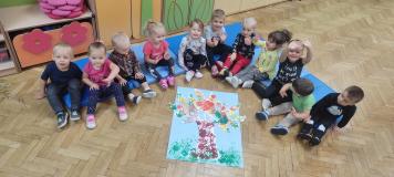 Grupa dzieci siedząca w półokręgu z wykonaną przez siebie pracą zbiorową przedstawiającą drzewo