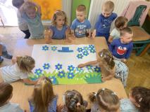 Zdjęcie przedstawiające grupę dzieci przy stoliku. Wspólnie wykonują pracę plastyczną przedstawiającą zieloną trawę i kwitnące niebieskie niezapominajki.